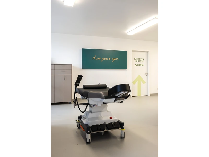 Patientenliege im hellen Flur vor petrolfarbenem Wandschild
